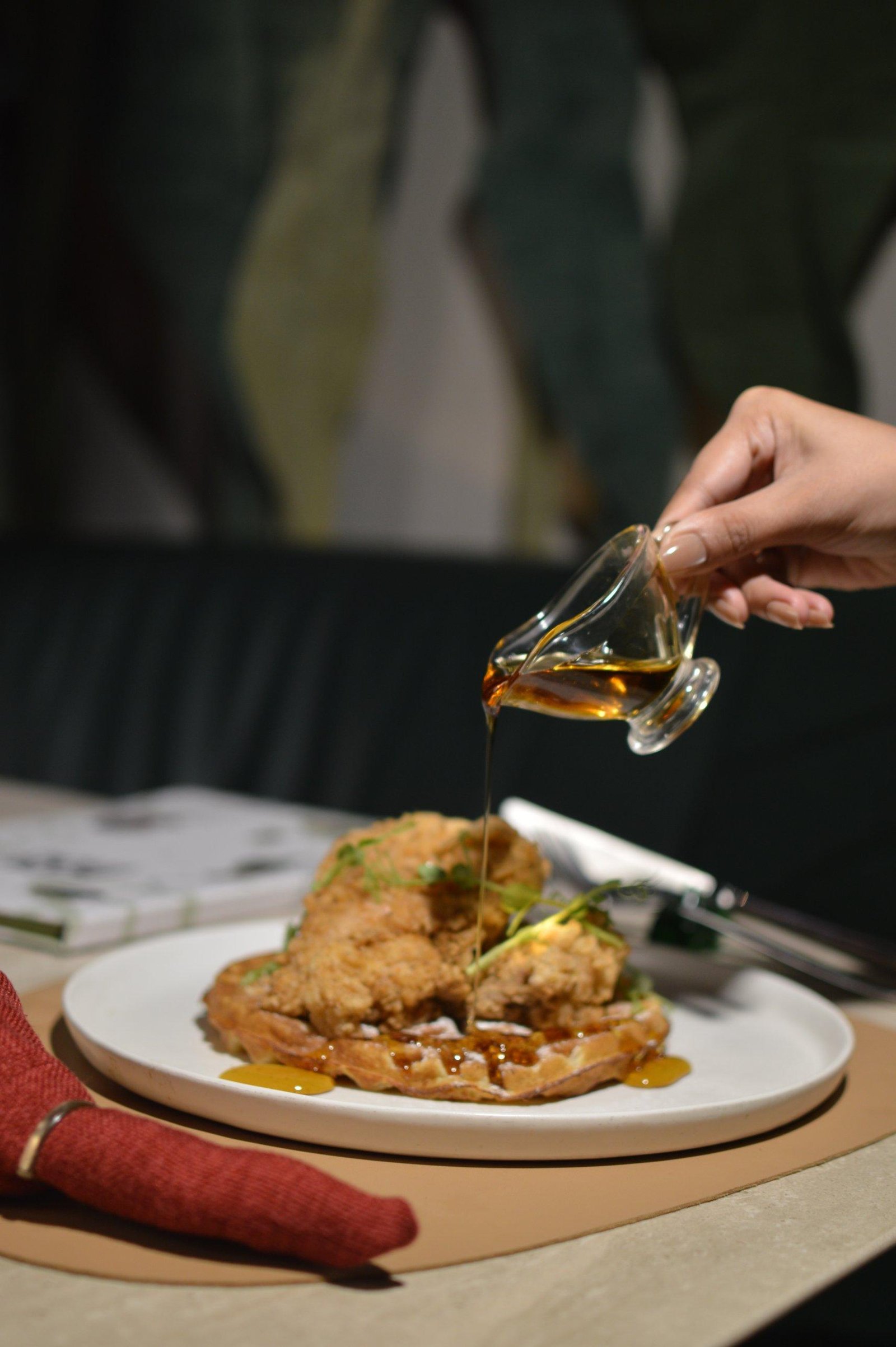 A hand pouring syrup over fried chicken and waffles at the table