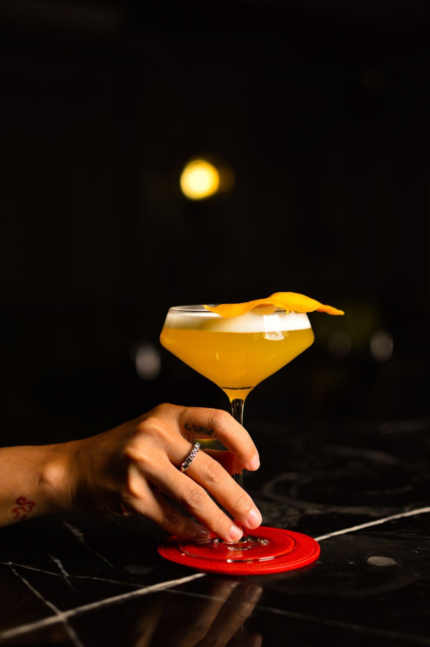 A hand holding a golden cocktail on a red coaster at a dark marble bar